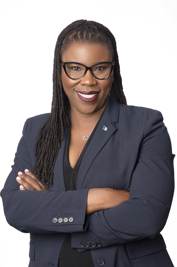 A woman with long, braided hair wearing glasses and a navy blazer stands smiling with her arms crossed against a white background.